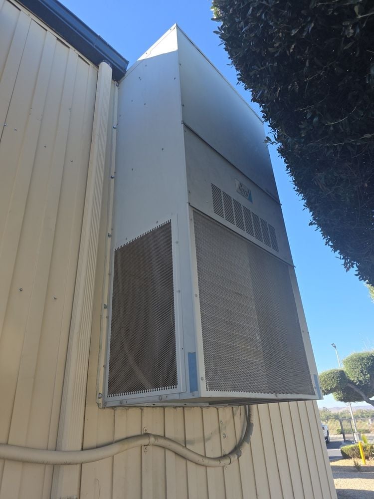 Large metal industrial air conditioning or HVAC unit mounted on corrugated metal building exterior against clear blue sky